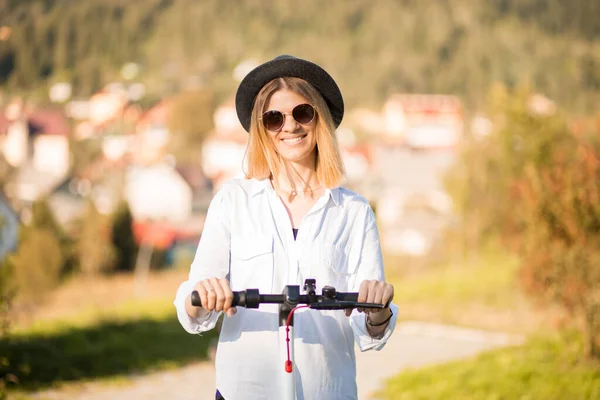 Trendy unbeschwerte Frau in weißem Hemd und schwarzem Hut fahren auf Tretroller Outdoor in der sommerlichen Parkstraße. Ökologisches Elektroverkehrskonzept. — Stockfoto