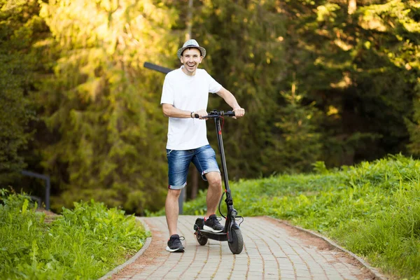 전기 운송. 하얀 셔츠를 입고 선글라스를 쓰고 모자를 쓴 젊은 남자가 맑은 여름에 시내 거리에서 전기 스쿠터 자전거를 탈준비를 하고 있습니다. 생태학적 운송 개념. — 스톡 사진