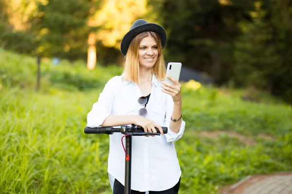 Retrato al aire libre de la joven hermosa niña en sombrero negro de pie con su scooter eléctrico mientras escribe en su teléfono. Árboles y colores verdes en el fondo. — Foto de Stock