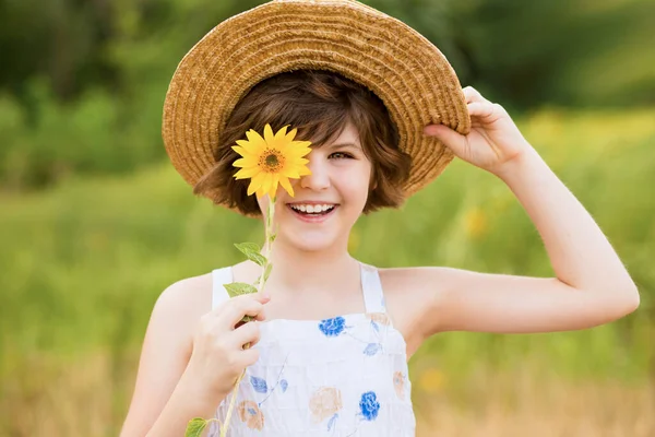 Belle petite fille en chapeau de paille avec des cheveux flottants cachent oeil avec fleur de tournesol, marche en plein air pendant les vacances d'été — Photo