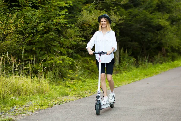 Vidám, gyönyörű lány fekete kalapban és fehér ingben, elektromos robogóval a nyári parkban. Scooter kölcsönzés, a mozgás szabadsága — Stock Fotó