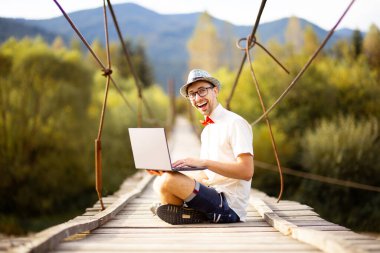 Gözlüklü mutlu genç serbest yazar dağ nehri üzerindeki köprüde dizüstü bilgisayarla oturuyor. İnternetteki web sörfü bilgileri ya da çevrimiçi çalışma