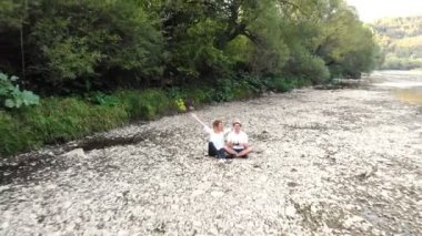 Nehir kenarında oturan çiftin havadan çekilmiş görüntülerini çekin ve görkemli doğa manzarasının tadını çıkarın. Dağlarda gezen insanlar, panoramik manzara, seyahat ve özgürlük, insansız hava aracı uçuşları.. 