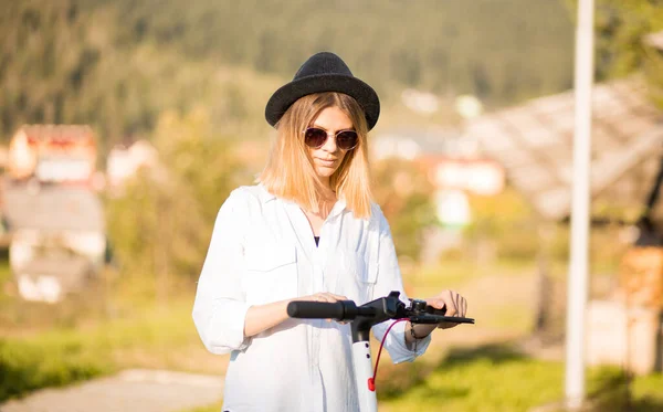 Young blond woman in black hat and sunglasses sets ride mode on touch panel. Stylish girl riding on electric scooter in city center. Summer leisure activity. Spending free time outdoors in sunny