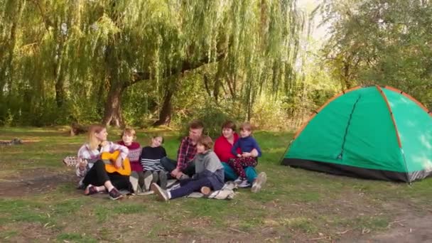 Grupp glada människor med barn sjunger samtidigt som du kopplar av på sommarsemester på campingtur. Kvinna spelar akustisk gitarr medan vänner njuter av fritiden — Stockvideo