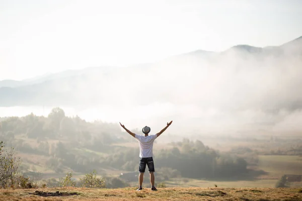 Happy traveler man raising hands up on top of world above mountains in white clouds. Hipster guy enjoy amazing atmospheric moment. travel and wanderlust concept