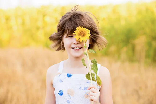 Menina bonito vestir vestido com girassol no campo de verão. Menina feliz esconder o olho com girassol. Férias de verão conceito. — Fotografia de Stock