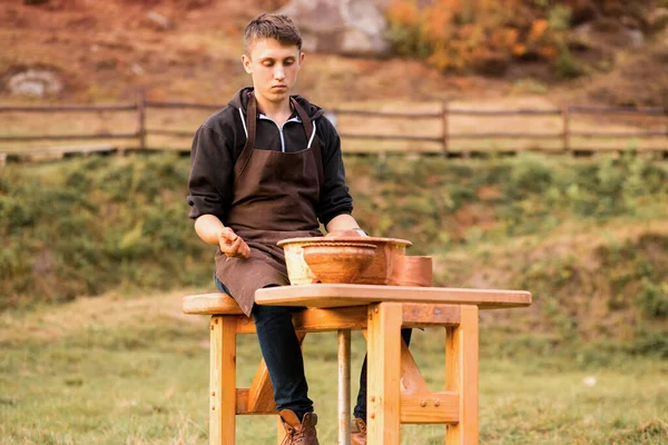 Молодой человек в фартуке сидит за гончарным колесом, формируя влажную глину на вращающемся поворотном столе летом на открытом воздухе — стоковое фото