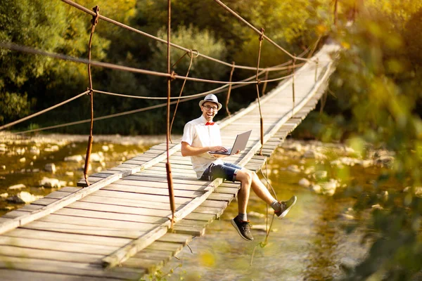 Inteligente guapo macho en sombrero, camisa blanca y gafas se sientan con las piernas cruzadas y sostienen el ordenador portátil. El hombre trabaja al aire libre con el ordenador portátil sentado en el bosque de montañas. Concepto de trabajo remoto y freelancer — Foto de Stock