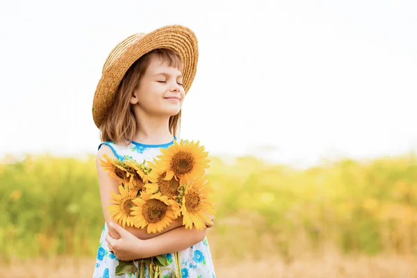 Retrato ao ar livre de adorável menina em chapéu de palha e vestido azul está andando em um campo de girassóis, reunindo e coletando buquê. Férias de verão — Fotografia de Stock