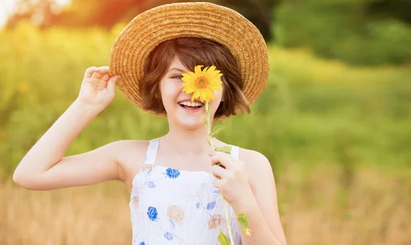 Belle petite fille en chapeau de paille avec des cheveux flottants cachent oeil avec fleur de tournesol, marche en plein air pendant les vacances d'été — Photo