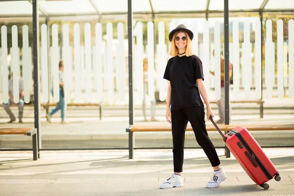 Jeune voyageuse occasionnelle en noir total et lunettes de soleil marchant et tirant des bagages à roues rouges. Concept de voyage. Espace de copie — Photo