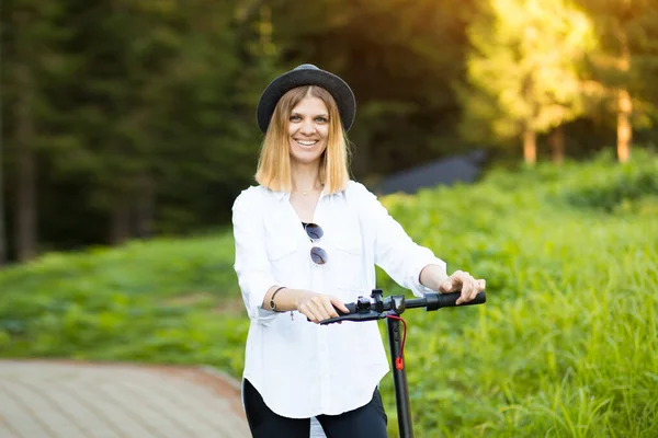 Trendy unbeschwerte Frau in weißem Hemd und schwarzem Hut fahren auf Tretroller Outdoor in der sommerlichen Parkstraße. Ökologisches Elektroverkehrskonzept. — Stockfoto