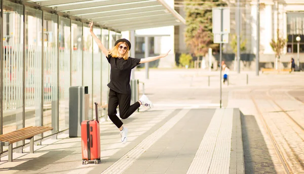 Femme touriste excitée porter noir total avec des bagages sautant sur la station de tram. Fille joyeuse voyageur saut avec valise. Concept de voyage. Profitez de frontières ouvertes pays sûr laissant — Photo