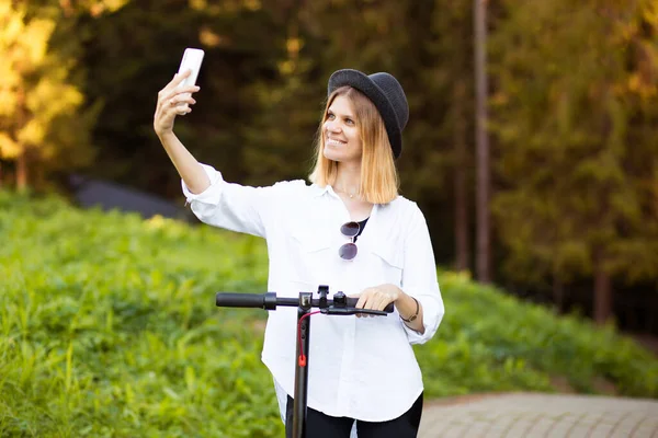 Retrato al aire libre de la joven y hermosa blogger con sombrero negro de pie con su scooter eléctrico y tomar selfie en su teléfono. Árboles y en el fondo. — Foto de Stock