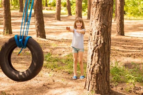 Cute little girl swinging na kole dołączony do dużego drzewa w lesie. — Zdjęcie stockowe