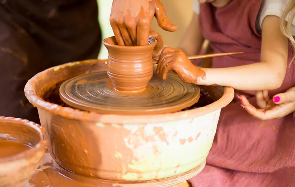 아이들을 위한 마스터 수업. 교사들의 손은 아이들에게 도공의 바퀴 위에서 도자기 요리를 만드는 방법을 보여 줍니다. 예술가는 진흙을 가지고 작업 합니다. 작은 소녀가 컵에 진흙을 조각하는 모습 — 스톡 사진