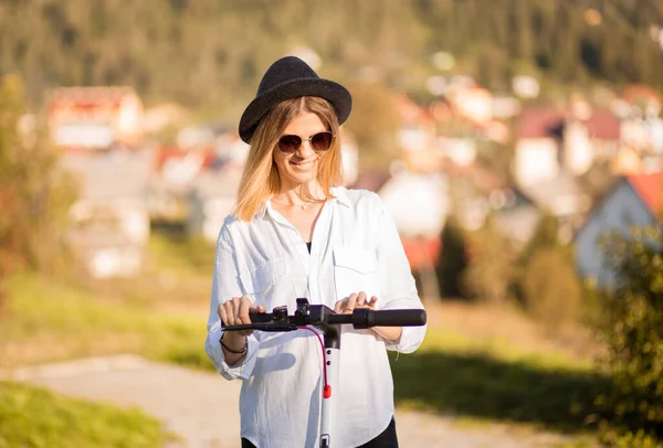 Mujer rubia joven en sombrero negro y gafas de sol establece el modo de paseo en el panel táctil. Elegante chica montando en scooter eléctrico en el centro de la ciudad. Actividad de verano. Pasar tiempo libre al aire libre en soleado —  Fotos de Stock