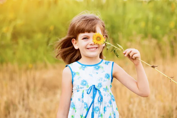 Linda niña usar vestido con girasol en el campo de verano. Feliz niña esconde el ojo con girasol. Concepto vacaciones de verano. —  Fotos de Stock