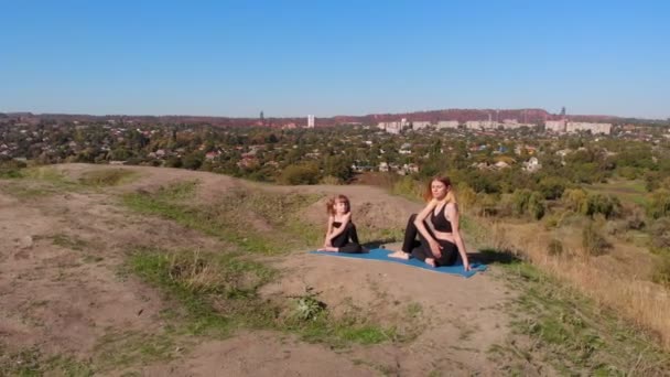 Vue aérienne de drone de mince jeune mère faire des exercices de yoga avec fille enfant sur la haute colline au bord du lac le matin, loin de l'agitation de la ville, donner le bon exemple et inculquer dans ses habitudes saines — Video