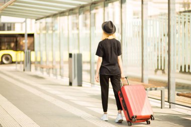 Siyah ve güneş gözlüklü genç bir bayan yolcu kırmızı tekerlekli bavullarını çekiyor ve yürüyor. Seyahat kavramı. Boşluğu kopyala
