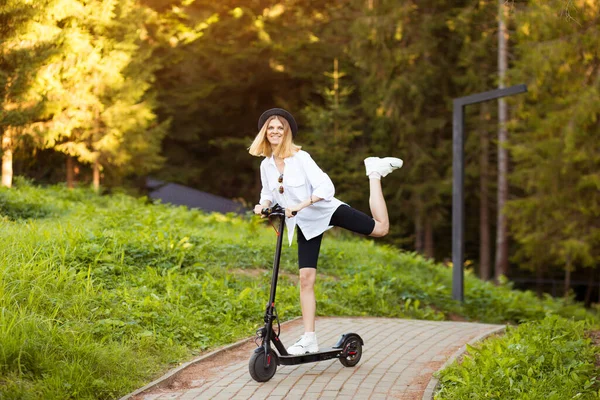 Sidovy av ung blond kvinna i vitt gå till jobbet på elektrisk skoter i staden sommar. — Stockfoto