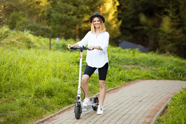 Vidám, gyönyörű lány fekete kalapban és fehér ingben, elektromos robogóval a nyári parkban. Scooter kölcsönzés, a mozgás szabadsága — Stock Fotó