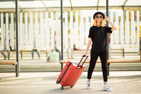 Jeune voyageuse occasionnelle en noir total et lunettes de soleil marchant et tirant des bagages à roues rouges. Concept de voyage. Espace de copie — Photo