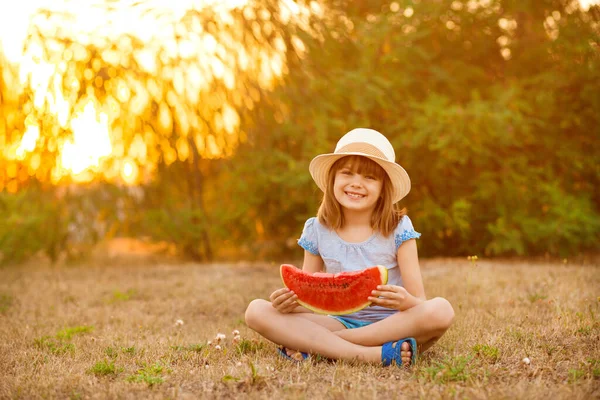 Adorable niña en sombrero de paja sentarse con las piernas cruzadas en la hierba verde, y come con entusiasmo jugosa sandía con sol en el fondo —  Fotos de Stock