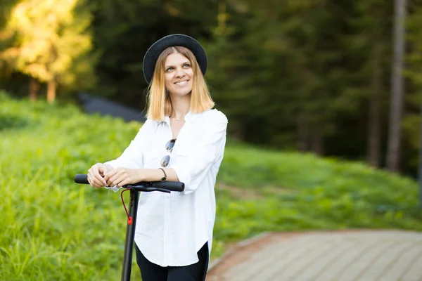 Trendy unbeschwerte Frau in weißem Hemd und schwarzem Hut fahren auf Tretroller Outdoor in der sommerlichen Parkstraße. Ökologisches Elektroverkehrskonzept. — Stockfoto