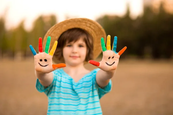 Glücklich lächelndes kleines Mädchen mit in lustiges Gesicht gemalten Händen, die draußen im Park spielen. Hände im Fokus — Stockfoto