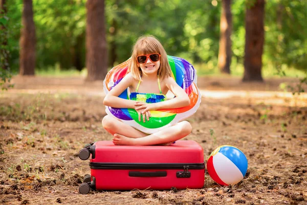 Traveler bambina si siede con le gambe incrociate sulla valigia in occhiali da sole e costume da bagno, pronto a volare al mare e avere vacanze attive — Foto Stock