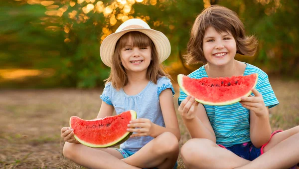 Due bambini sorridenti sorridenti siedono con le gambe incrociate sull'erba e mangiano anguria al parco estivo con tramonto — Foto Stock