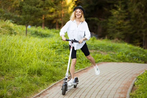 Vidám, gyönyörű lány fekete kalapban és fehér ingben, elektromos robogóval a nyári parkban. Scooter kölcsönzés, a mozgás szabadsága — Stock Fotó