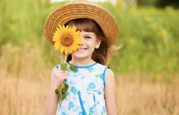 Красива маленька дівчинка в солом'яному капелюсі з рум'яною шерстю приховує око з квітками соняшнику, гуляючи на відкритому повітрі в літні канікули Стокове Зображення