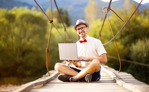 Inteligentny przystojny mężczyzna w kapeluszu, białej koszuli i okularach siedzieć ze skrzyżowanymi nogami i trzymać laptopa. Człowiek pracuje na zewnątrz z laptopem siedzącym w górach w lesie. Pojęcie pracy zdalnej i freelancera — Zdjęcie stockowe