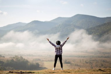 Mutlu kız kollarını kaldırıp dağların tepesinde duruyor ve muhteşem manzaranın tadını çıkarıyor. Beyaz bulutlarla kaplı yaz dağının tepesindeki siluet kadın yürüyüşçü..