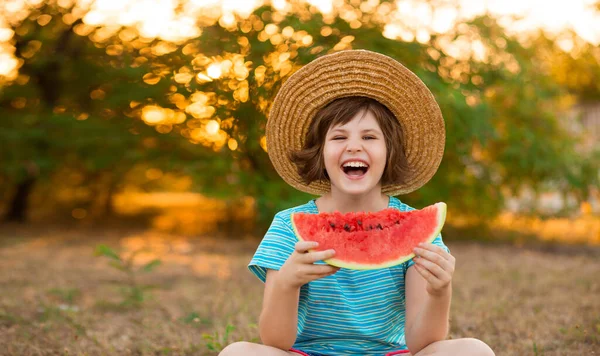 Adorable petite fille en chapeau de paille assis avec les jambes croisées sur l'herbe verte, et mange avec impatience pastèque juteuse avec soleil sur le fond — Photo