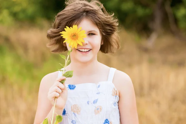 Menina bonito vestir vestido com girassol no campo de verão. Menina feliz esconder o olho com girassol. Férias de verão conceito. — Fotografia de Stock