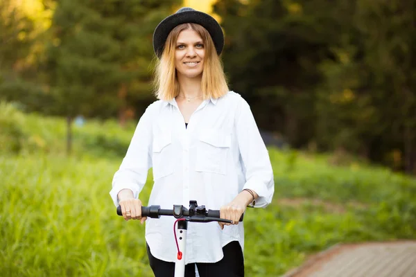 Trendy gondtalan nő fehér ingben és fekete kalap lovagolni kick robogó szabadban a Summer Park utcában. Elektromos ökológiai közlekedés koncepciója. — Stock Fotó