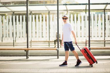 Tren istasyonunda güneş gözlüklü, sakallı, kırmızı valizli yakışıklı bir turist. Seyahat ve tatil kavramı. Yolculuk. Boşluğu kopyala. 