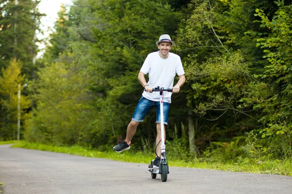 Elektrisk transportering.Ung stilig man i vit skjorta, solglasögon och hatt redo att rida sin elektriska skoter cykel på stadens gata i solig sommar. Begreppet ekologiska transporter. — Stockfoto