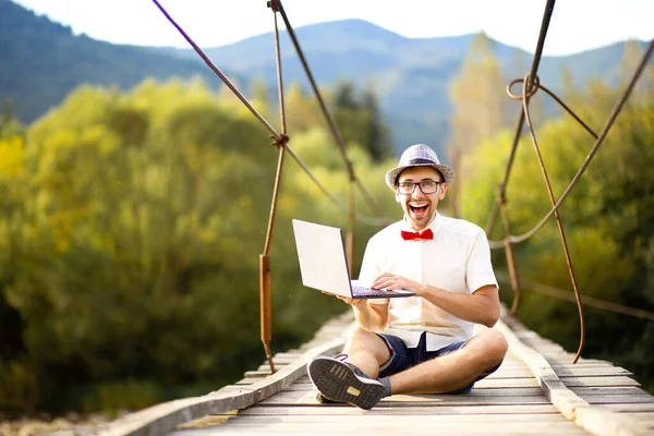 Intelligente bel maschio in cappello, camicia bianca e occhiali sedersi con le gambe incrociate e tenere il computer portatile. L'uomo lavora all'aperto con il computer portatile seduto nella foresta di montagne. Concetto di lavoro a distanza e freelance Immagini Stock Royalty Free