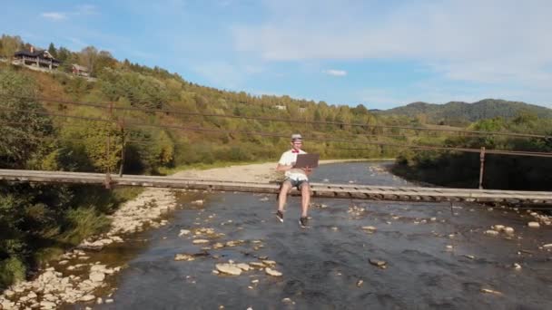 Drohnenbild eines jungen Mannes im weißen Hemd, der auf einer Holzbrücke über den Gebirgsfluss sitzt und Laptop benutzt. — Stockvideo
