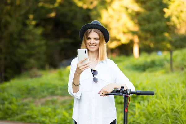 Retrato al aire libre de la joven hermosa niña en sombrero negro de pie con su scooter eléctrico mientras escribe en su teléfono. Árboles y colores verdes en el fondo. — Foto de Stock