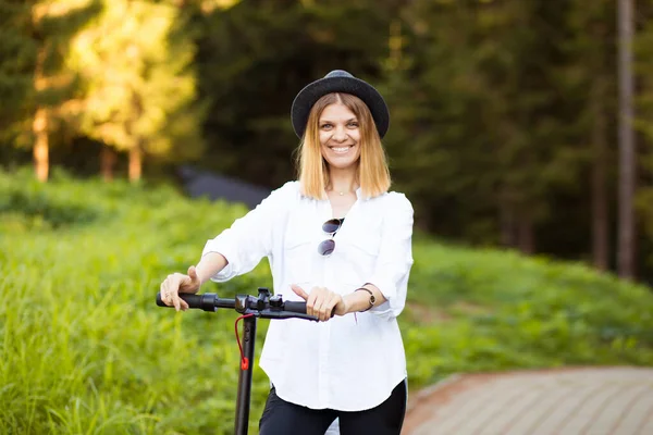 Trendy unbeschwerte Frau in weißem Hemd und schwarzem Hut fahren auf Tretroller Outdoor in der sommerlichen Parkstraße. Ökologisches Elektroverkehrskonzept. — Stockfoto