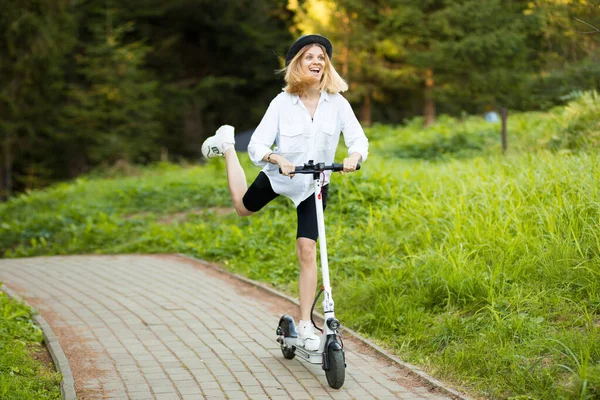 夏の公園で電動スクーターに乗って黒の帽子と白いシャツの陽気な美しい女の子。スクーターのレンタル、動きの自由 — ストック写真