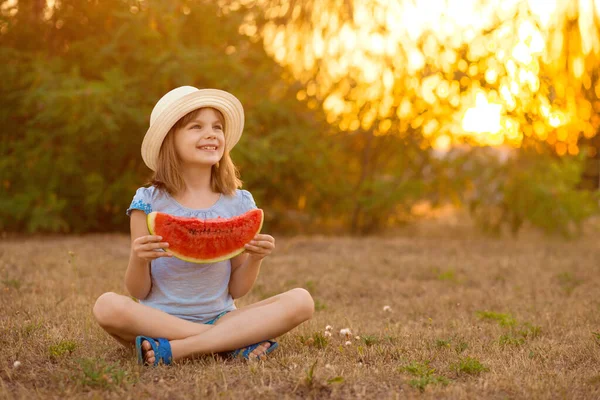 Adorable niña en sombrero de paja sentarse con las piernas cruzadas en la hierba verde, y come con entusiasmo jugosa sandía con sol en el fondo —  Fotos de Stock