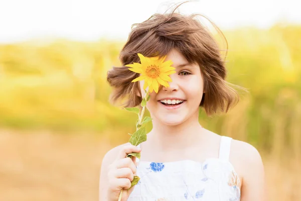 Menina bonito vestir vestido com girassol no campo de verão. Menina feliz esconder o olho com girassol. Férias de verão conceito. — Fotografia de Stock