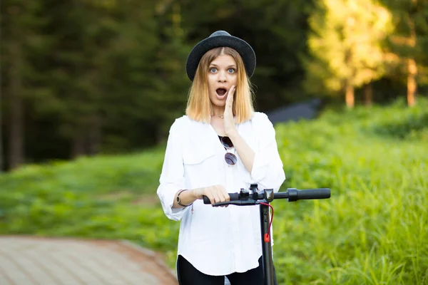 Trendy gondtalan nő meglepett kifejezés fehér ing és fekete kalap lovagolni kick robogó szabadban nyári park utcában. Elektromos ökológiai közlekedés koncepciója. — Stock Fotó